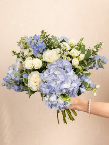 Enora bouquet being held by a woman.  Mixed coastal bouquet featuring blue hydrangeas