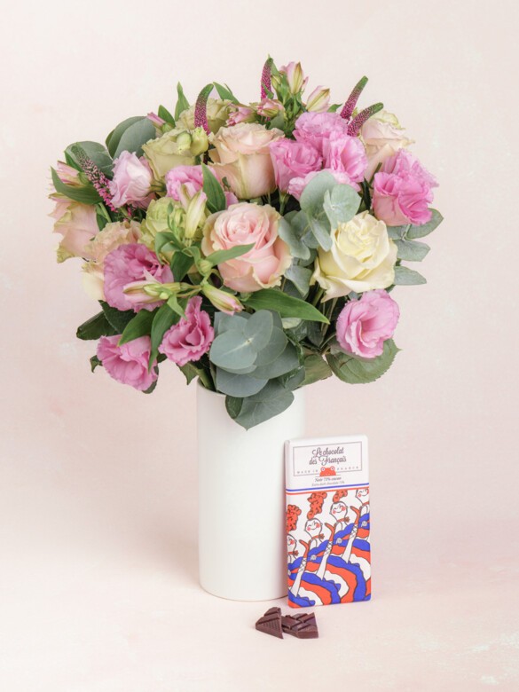 A bouquet of soft pink and ivory flowers in a tall white vase, paired with an artisanal French chocolate bar and broken dark chocolate pieces on display.