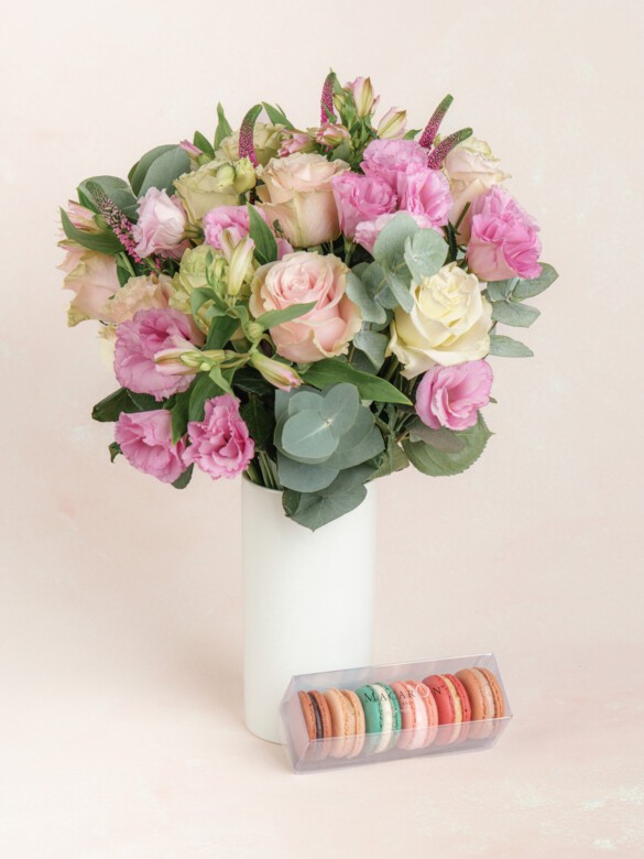 A tall white vase holding a bouquet of pink and ivory flowers, displayed alongside a clear box of colorful assorted macarons.