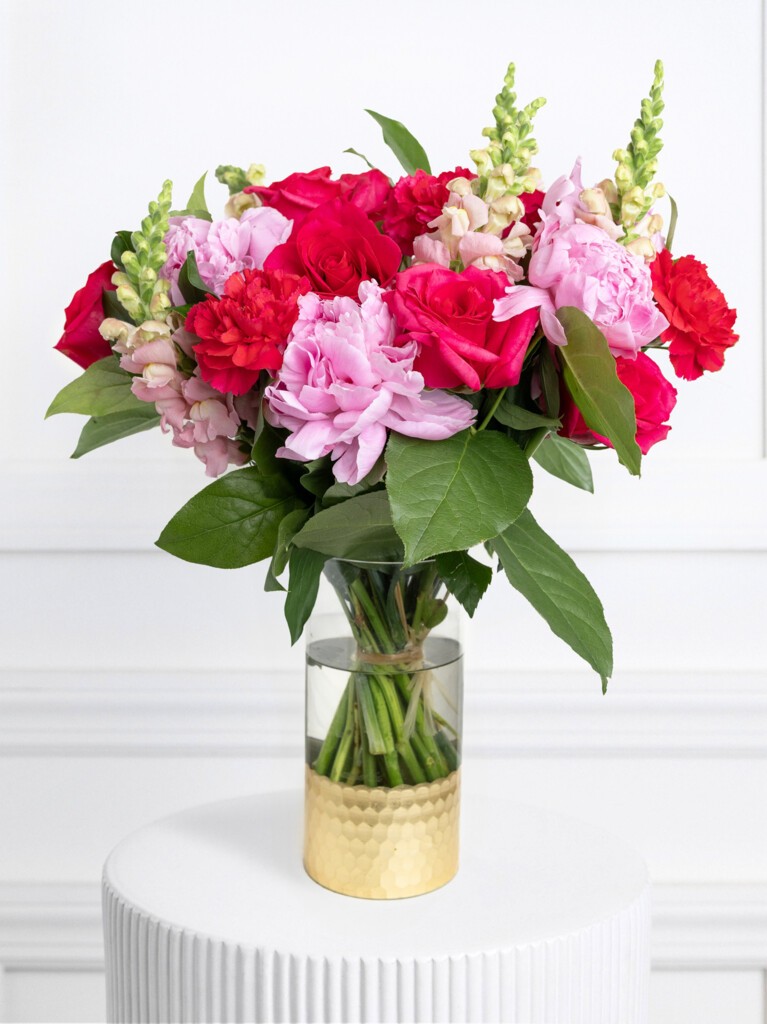 A vibrant bouquet of 26 stems arranged in a clear glass vase with a golden textured base, blending deep red roses, fiery red carnations, and plush pink peonies with pale snapdragons and lush green foliage for a dramatic, regal look.