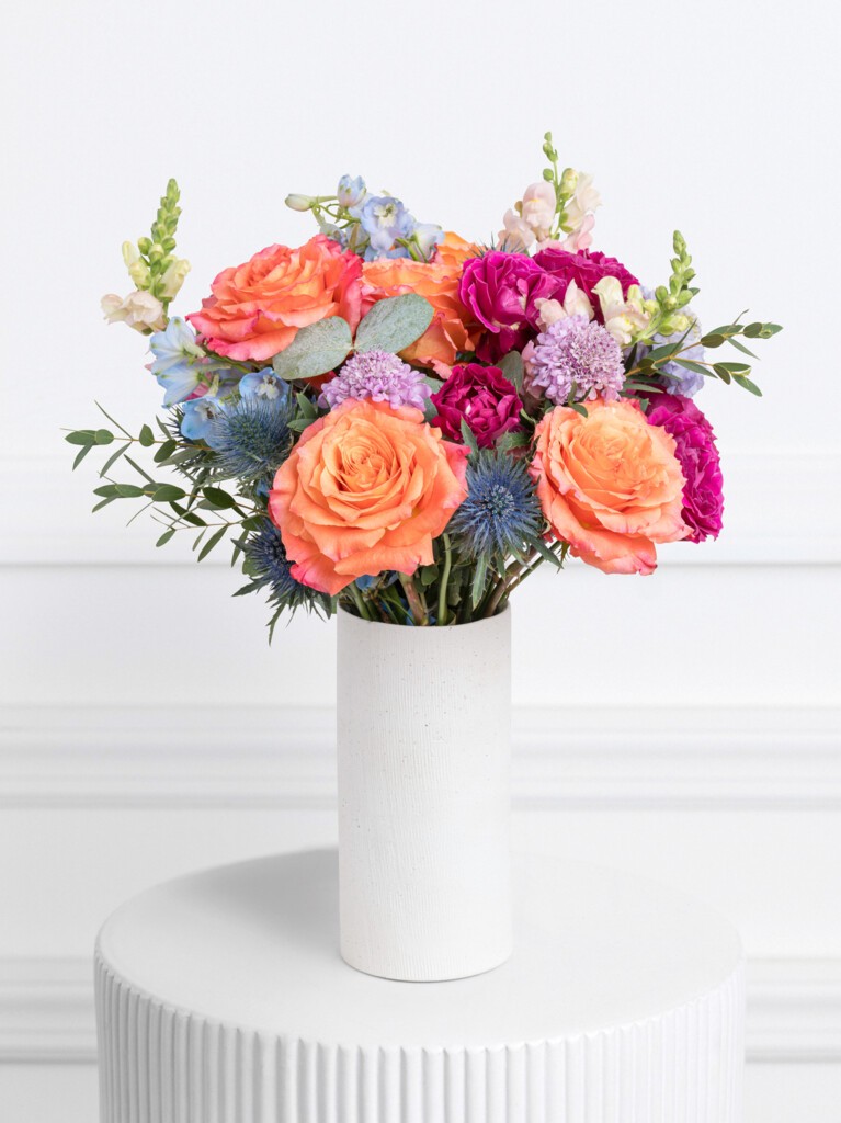 A vivid floral arrangement bursting with orange roses, magenta carnations, soft snapdragons, lavender scabiosa, thistle, and blue delphinium, all styled in a tall white ceramic vase.