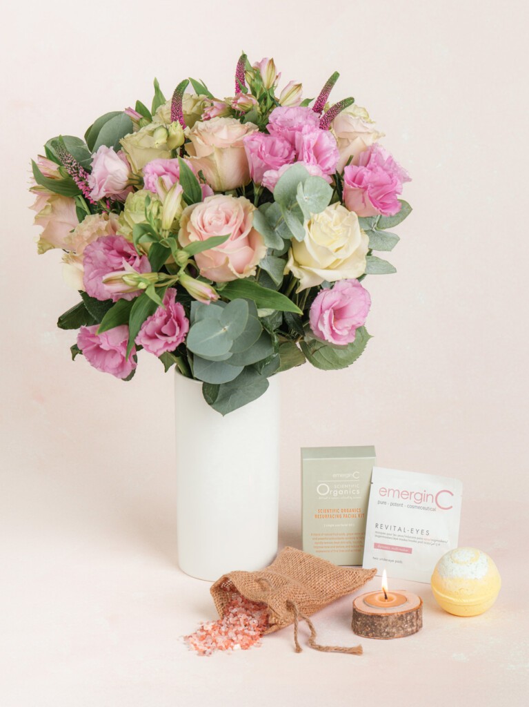 A pink and white floral bouquet in a tall white vase, paired with a spa kit featuring a rustic candle, bath bomb, and skincare essentials, all arranged on a linen produce bag.