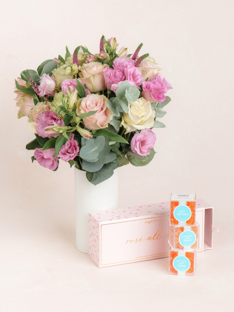 A bouquet of pink and cream flowers in a tall white vase, displayed alongside a stacked trio of Sugarfina gummy candy cubes in rose-toned packaging.