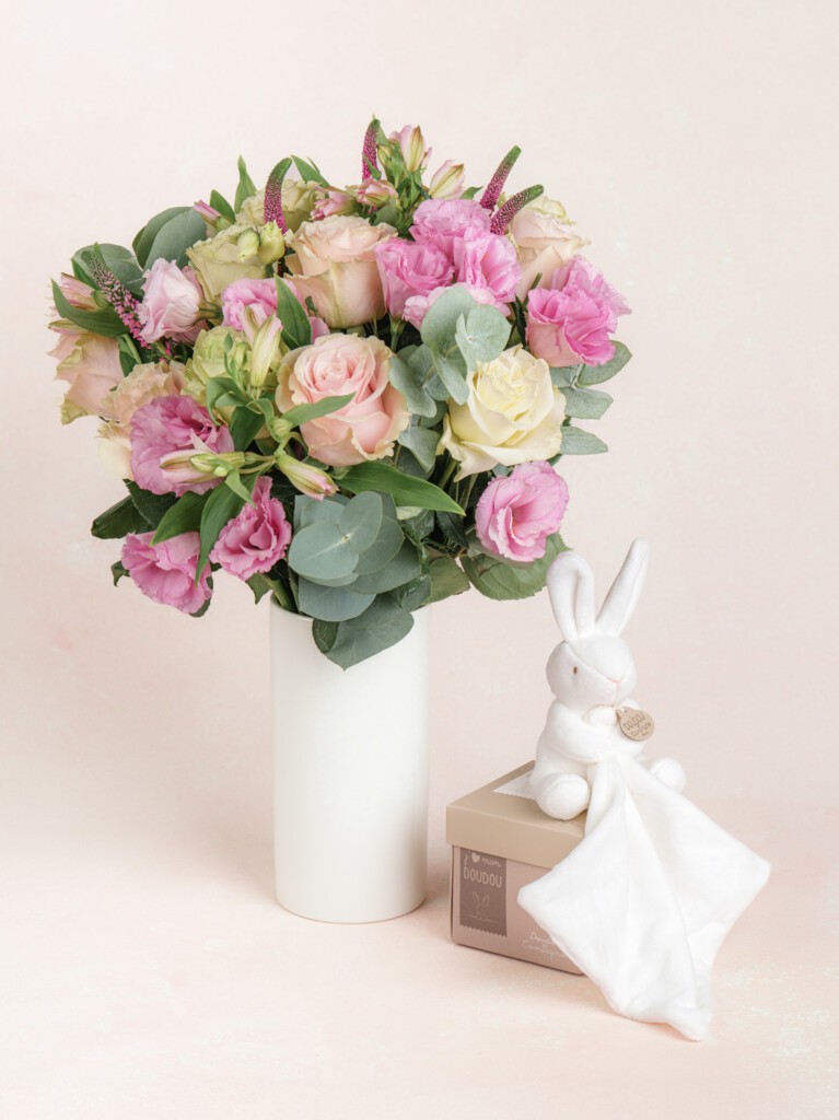 A bouquet of soft pink and ivory flowers in a tall white vase, paired with a white rabbit clutching a cream satin-trimmed baby blanket.