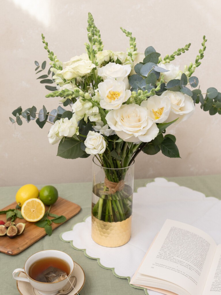 Paulette bouquet with 26 white blooms and eucalyptus displayed on a table beside tea and citrus. A serene floral accent for elegant mornings or thoughtful gifting.