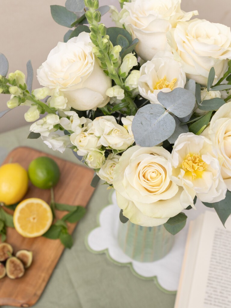 Top view of Paulette 16-stem bouquet with white roses, snapdragons, and eucalyptus, styled on a table with citrus and figs for a fresh home accent.