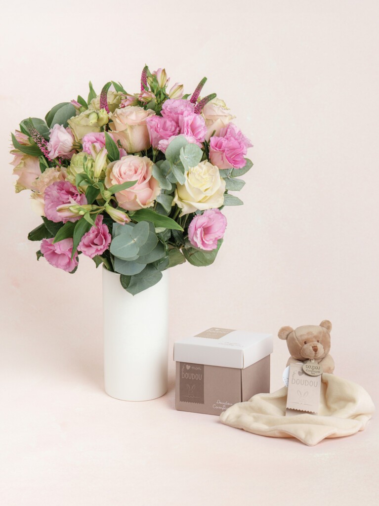 A bouquet of soft pink and ivory flowers in a tall white vase, paired with a teddy bear clutching a cream satin-trimmed baby blanket.