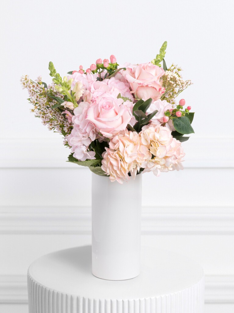 Petite bouquet in a tall white vase, combining blush pink Mondial roses, pale hydrangeas, and delicate sprigs of snapdragon, waxflower, and hypericum, accented with soft greenery and rice flower for a textured, airy pastel arrangement.