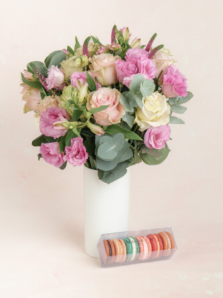 A tall white vase holding a bouquet of pink and ivory flowers, displayed alongside a clear box of colorful assorted macarons.