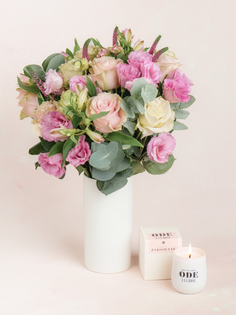 A bouquet of blush and pink flowers arranged in a tall white vase, paired with a lit white petite candle labeled “Ode à la Rose” placed in front.