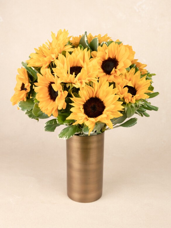 A full, round bouquet of bright yellow sunflowers with dark centers and lush green foliage, arranged in a tall, brushed gold cylindrical vase.