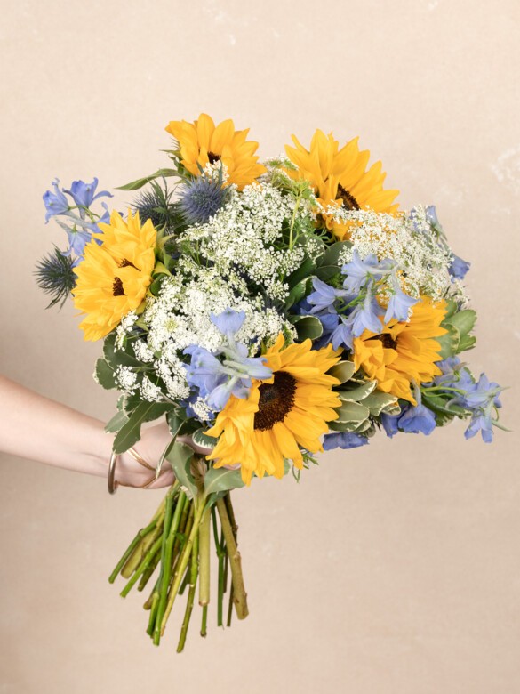 A hand-tied bouquet of vibrant sunflowers, delicate Queen Anne’s lace, blue delphinium, and thistle, creating a fresh, summer-filled explosion of color.